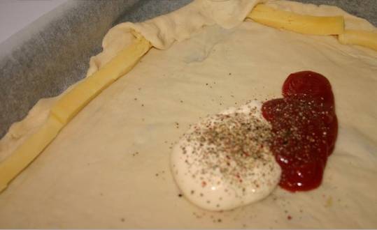 1. Тесто разморозим, раскатаем в тонкий пласт. Сделаем вилкой несколько отверстий, чтобы тесто не "пузырилось" при выпекании. Формируем бортики. Основу смажем майонезом и томатной пастой, а сыр выкладываем брусочками и как бы "заворачиваем" в бортики. Посыпаем основу специями. 