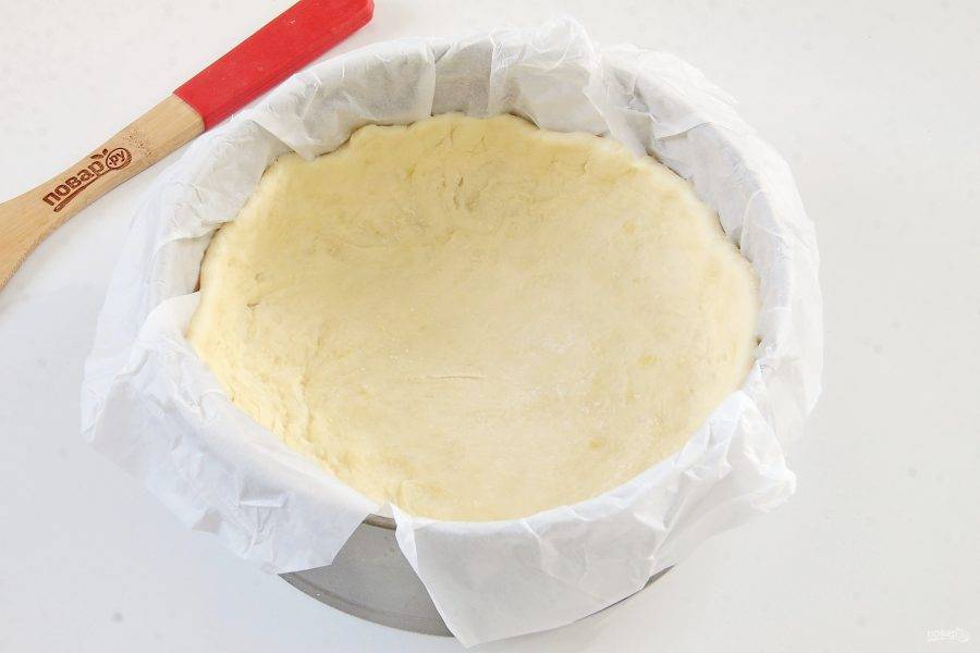 Разъемную форму, диаметром не более 20 см, застелите пергаментом, смажьте маслом и положите тесто, равномерно распределив его по всей форме. Сформируйте высокие бортики. Если необходимо, посыпьте тесто мукой.