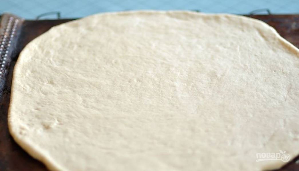 Возьмите дрожжевое тесто для пиццы, разморозьте его, если оно лежало в морозилке, раскатайте тесто в пласт около 0,5 сантиметра толщиной. Выложите тесто на противень. 