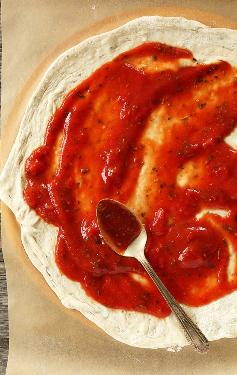 На противень положить бумагу для выпечки. Тесто для пиццы тонко раскатать на бумаге, томатный соус распределить равномерно по поверхности пиццы, посыпать специями.