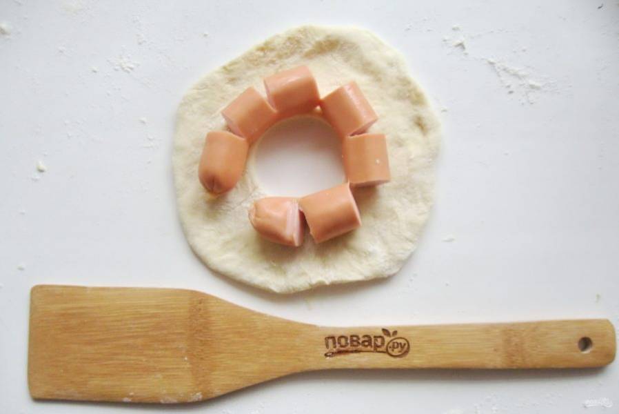 Тесто разделите на восемь частей. Каждую часть раскатайте в лепешку. Сосиску надрежьте  и выложите на лепешку, в середине которой сделайте дырочку, как у пончика.