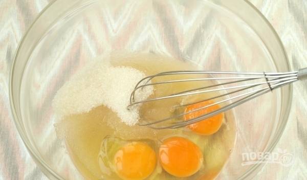 В стеклянную миску с высокими бортиками всыпьте сахар и вбейте сырые куриные яйца. Затем тщательно взбейте все венчиком до однородности.