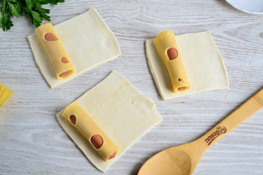 Каждый квадрат раскатайте в одну сторону, чтобы слегка удлинить его. На каждый кусок теста положите сосиску, вокруг которой оберните тонкий ломтик сыра. Я покупаю уже нарезанный сыр, с ним легче работать.