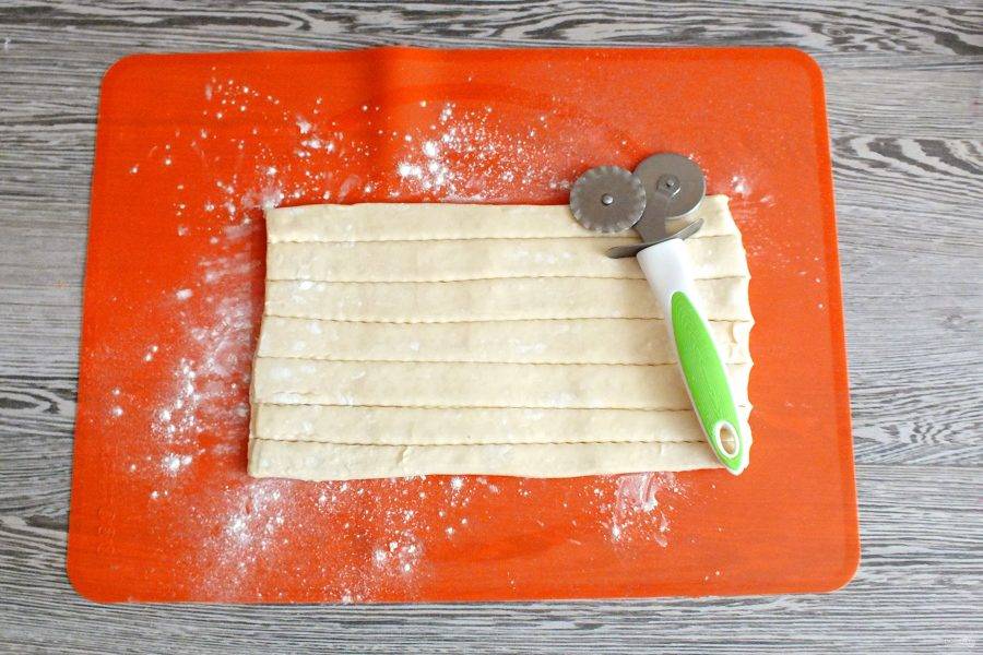 Заранее размороженное тесто раскатайте в одном направлении. Длина пласта должна быть 26-28 см. Нарежьте полоски шириной 2 см.