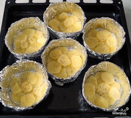 Формочки из фольги переложите на противень и смажьте маслом. Картофель помойте, почистите и нарежьте кружками. Разложите его по формам. Посолите и поперчите картофель.
