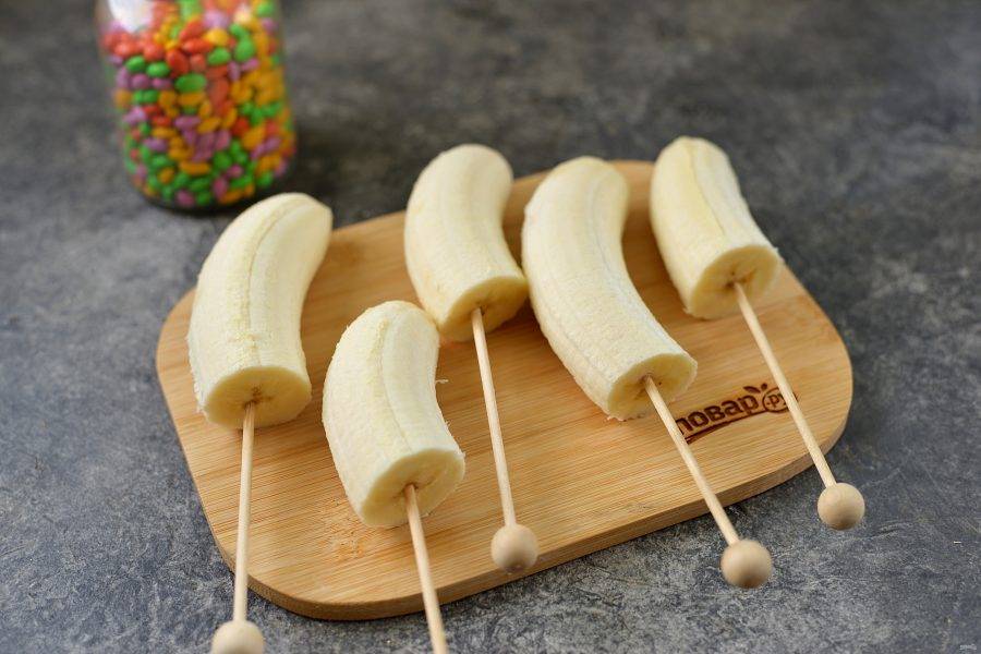 Почистите бананы, разрежьте их пополам, вставьте в каждый деревянную шпажку.