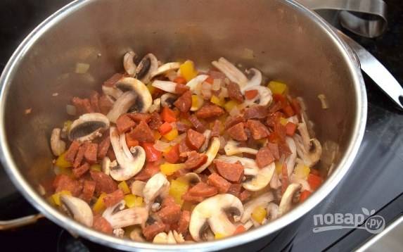Грибы помойте, нарежьте слайсами. Колбасу также нарежьте тонкими колечками. Добавьте грибы и колбасу к овощам и обжарьте еще 2-3 минуты.