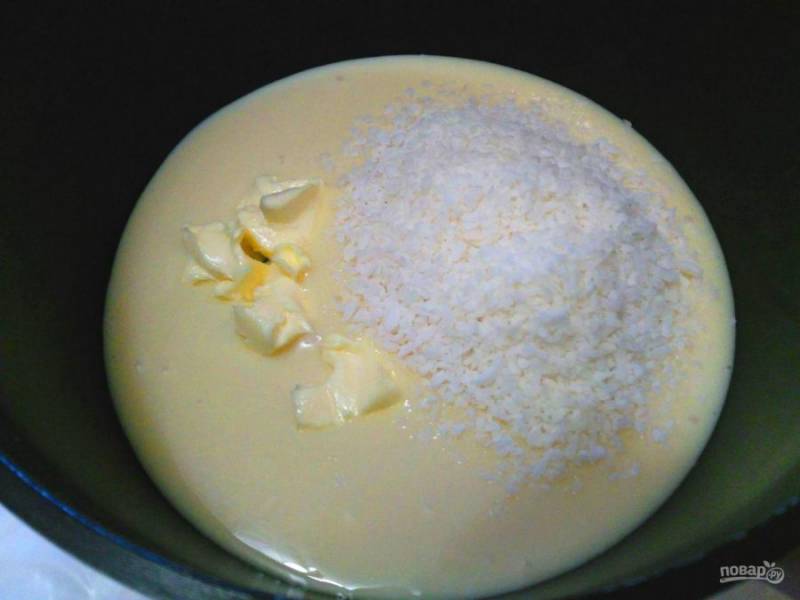 В толстостенную кастрюльку или сотейник вылейте сгущенное молоко, добавьте порезанное кубиками масло и кокосовую стружку.