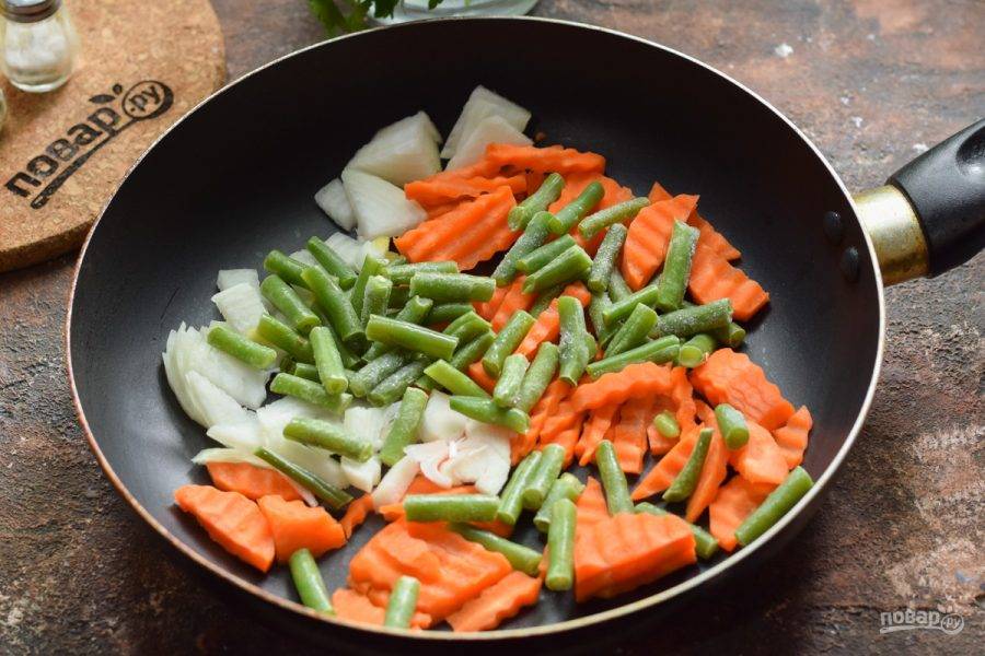 В сковороде прогрейте масло, выложите лук и морковь, добавьте стручковую фасоль.