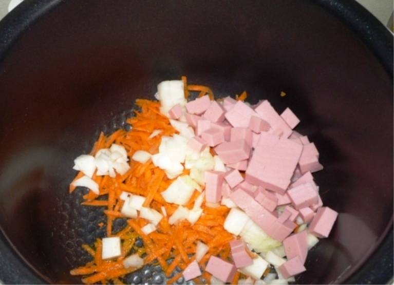 В чашу мультиварки налейте немного масла, положите тертую морковь, порезанные лук и колбасу. 