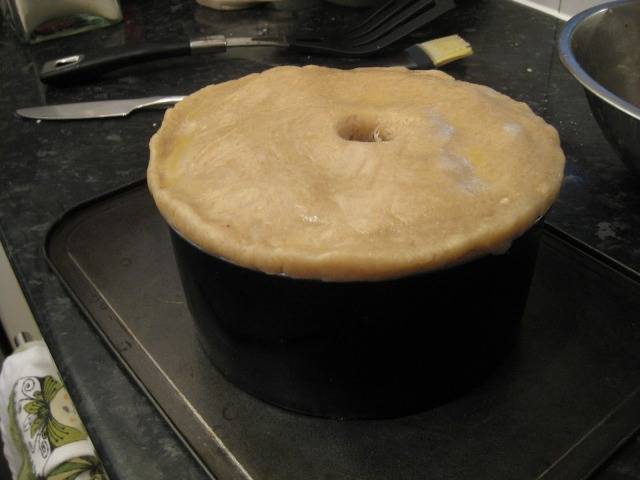 Из оставшегося теста формируем крышечку. Смазываем края пирога водой, чтобы крышечка к нему хорошо пристала, и защипываем. В центре крышечки делаем отверстие. Смазываем верх яйцом.