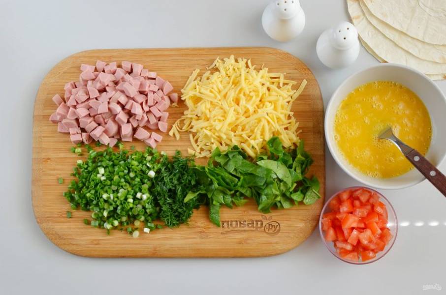 Взболтайте три яйца с солью и перцем. Натрите сыр на терке, порежьте кубиками сосиски, порежьте зелень. Помидор разрежьте на 4 части, удалите семена, а плотные части порежьте кубиками.