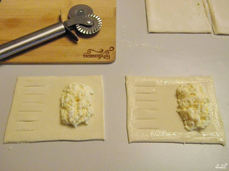 Порежьте тесто, не раскатывая на прямоугольники. С одной стороны сделайте красивые надрезы. Весь периметр смажьте белком, чтобы лучше склеился край слойки. В центр выложите по столовой ложке творога.