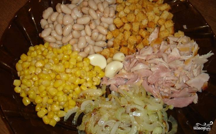 2. Открываем банки с кукурузой и фасолью, сливаем с них лишнюю жидкость. Фасоль откидываем на сито и немного промываем в теплой воде. Окорок очищаем, нарезаем мелко мясо. Чистим чеснок и измельчаем через пресс или натираем на терке.