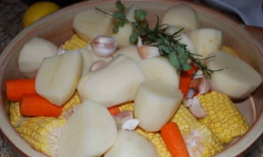 Противень смазываем растительным маслом, духовку прогреваем до 200 градусов. Крупно нарезаем картофель и морковь. Кукурузу режем на несколько частей. Лук нарезаем дольками, чеснок очищаем и разбираем. Выкладываем все овощи на противень.