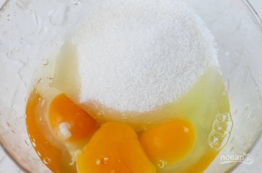 В отдельную миску вбейте сырые куриные яйца. Добавьте к ним соль и стакан сахара. Взбейте ингредиенты при помощи миксера или кухонного комбайна до появления плотной пены.  