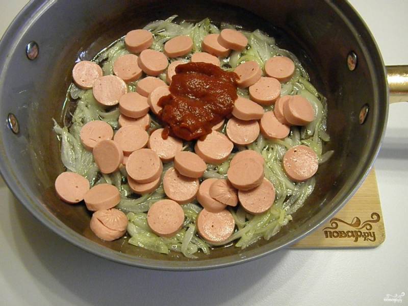 К полуготовому луку добавьте сосиски и томатную пасту, обжарьте пару минут. Добавьте квашенную капусту, перец черный и соль, если нужно. Перемешайте, влейте воду и протушите до мягкости под крышкой.