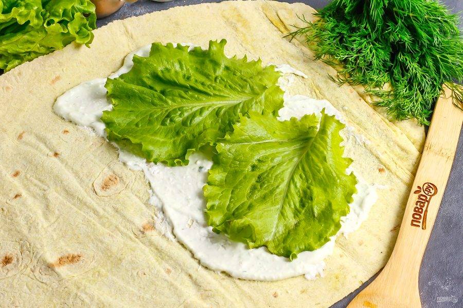 Разверните тонкий лаваш на рабочей поверхности, его край смажьте половиной сметанного соуса. Промойте листья салата, и удалите стебли. Выложите их на соус.