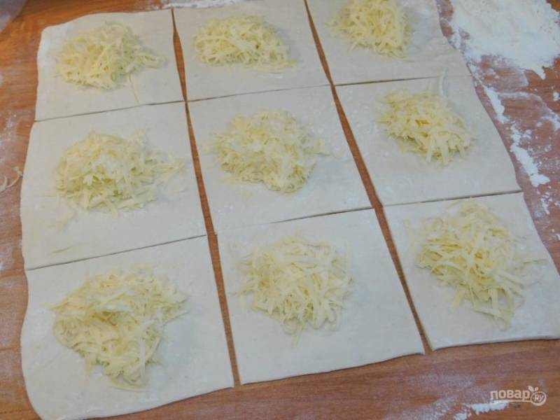 Затем рулет раскатайте еще раз в тонкий пласт теста. Нарежьте на квадраты и смажьте небольшим количеством растопленного масла. Выложите в центр тертый сыр. 
