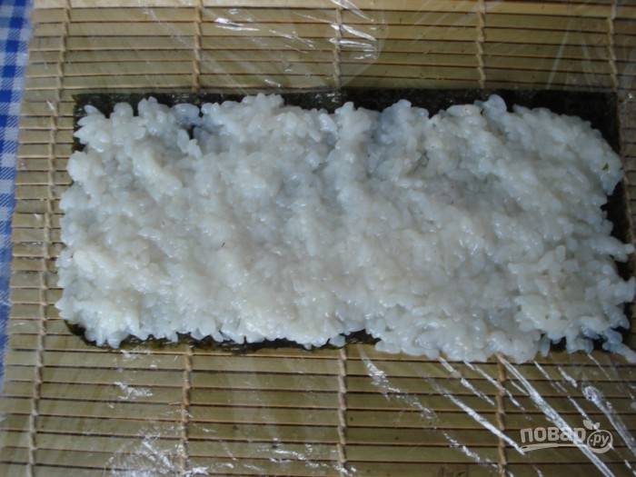 Выложите на стол бамбуковый коврик. Сверху пищевую плёнку. Разложите нори. Смоченными водой руками распределите рис.