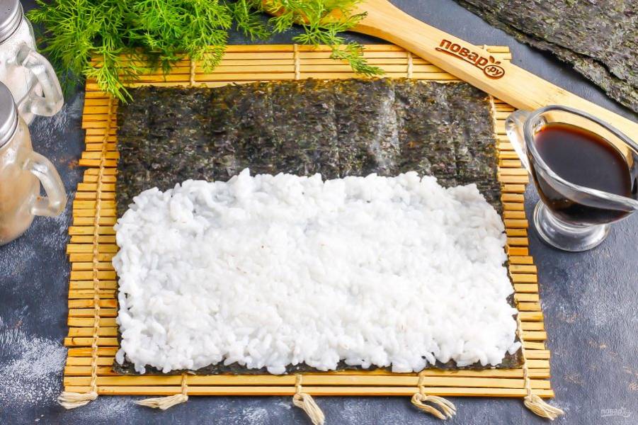 Выложите лист нори на коврик и распределите на него часть отварного риса, смочив руки в воде, иначе рис будет к ним прилипать. Оставьте незаполненным часть листа примерно в 2-3 см.