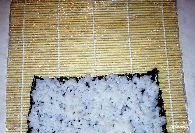 Расстелите циновку, обернутую в пищевую пленку. Таким образом она не будет загрязняться. На нее постелите лист нори. Выложите и равномерно распределите по нори рис. 