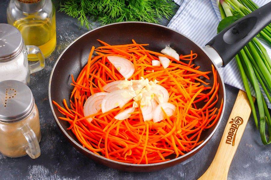 Нарежьте лук полукольцами. Влейте на сковороду растительное масло, добавьте нарезку моркови и лука, спрессуйте зубчики чеснока. Обжарьте примерно 2-3 минуты.