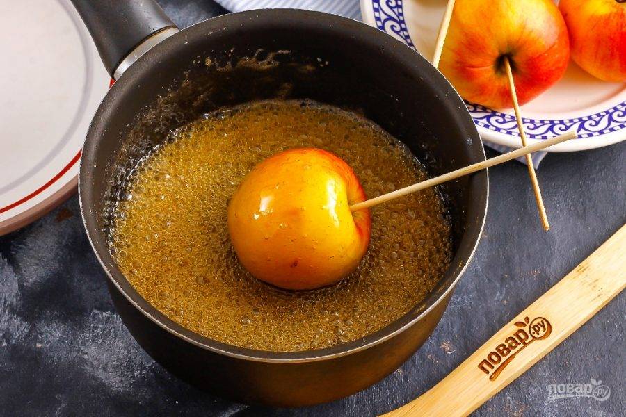 Снимите емкость с плиты и обмакните в нее каждое яблоко, чтобы карамель его покрыла полностью. Слегка наклоняйте емкость, чтобы сладость попала и в место скрепления яблока и шпажки.