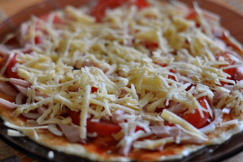 Теперь посыпаем стол мукой и выкладываем на него приготовленное тесто, раскатываем его в очень тонкий круглый пласт, чем он тоньше, тем быстрее приготовится пицца. Перекладываем нашу основу для пиццы на специальную плоскую тарелку для микроволновки, смазываем тесто томатной пастой и посыпаем специями. Выбранные для начинки продукты нарезаем небольшими кусочками и выкладываем их на пласт теста, солим немного, сверху посыпаем все обильно сыром.