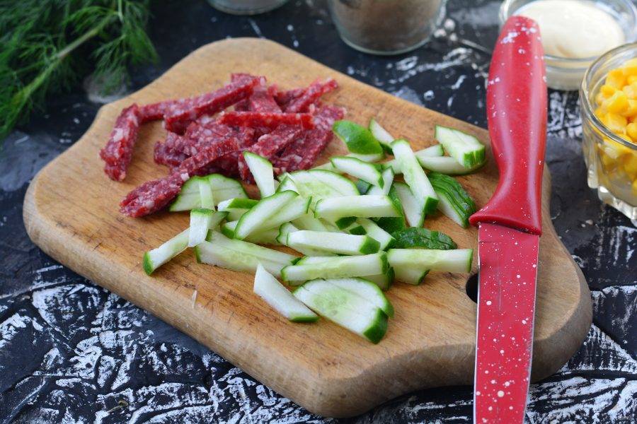 Нарежьте полосками копченую колбасу и свежие огурцы.