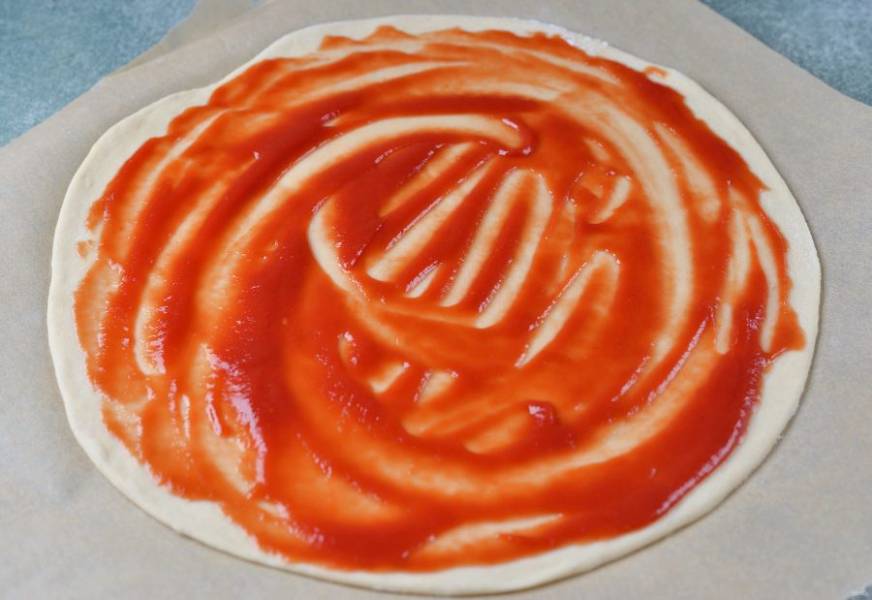 Подошедшее тесто нужно обмять и разделить на две части, у вас получится две пиццы диаметром 30 сантиметров. Тоненько раскатайте на пергаменте, при необходимости подсыпьте муки. Смажьте тесто любимым томатным соусом.