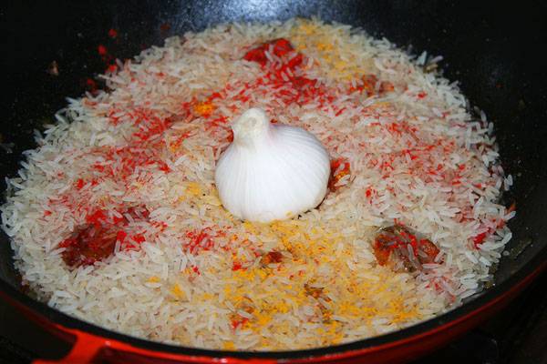 Засыпаем половину риса, еще немножко специй, затем вторую половину риса и снова специи. В самом верху кладем головку чеснока с обрезанной нижней частью. Заливаем блюдо кипятком, чтоб воды было на 1 см выше риса. Готовим на мелком огне, примерно 30 минут.