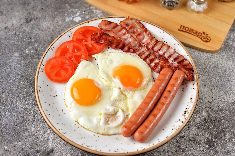 На большую плоскую тарелку выложите аккуратно яйца, сосиски и бекон, положите тонко нарезанный помидор.