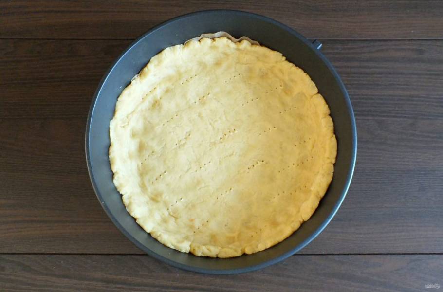 Возьмите форму для выпечки диаметром 26 см. Застелите ее пекарской бумагой. Выложите в форму тесто. Распределите его по дну, формуя высокие бортики. Основание пирога проткните вилкой. Поставьте в разогретую до 180 градусов духовку на 10 минут.