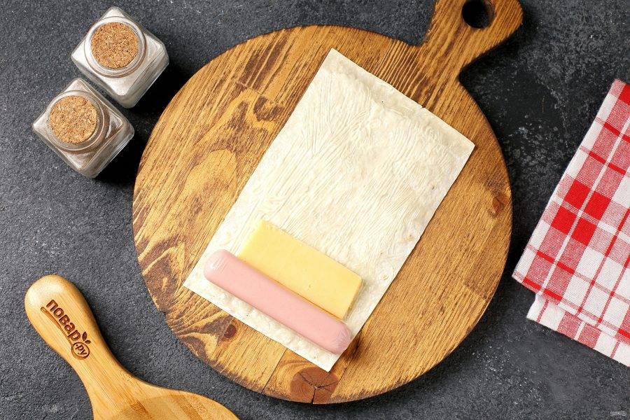 Смажьте каждую полоску лаваша тонким слоем майонеза, затем выложите на край сосиску и кусочек сыра, как на фото.