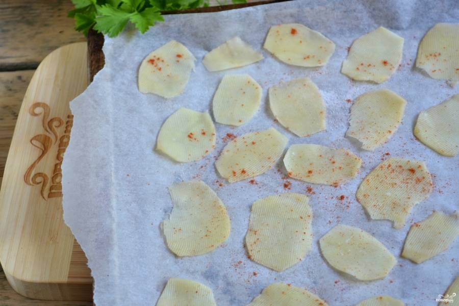 Выложите картофельные слайсы на пергамент, присыпьте их солью и паприкой, сбрызните подсолнечным маслом.