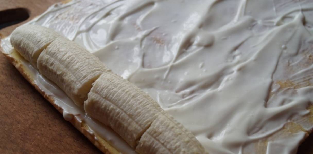 Аккуратно разверните бисквит. Равномерно распределите по нему крем. Выложите по краю (с которого начнёте сворачивать рулет) порезанный кусочками банан.