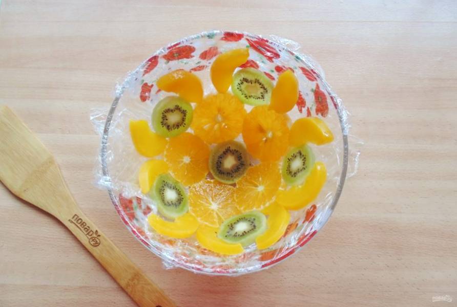 Миску застелите пищевой пленкой. Выложите нарезанный апельсин, киви и консервированные персики.