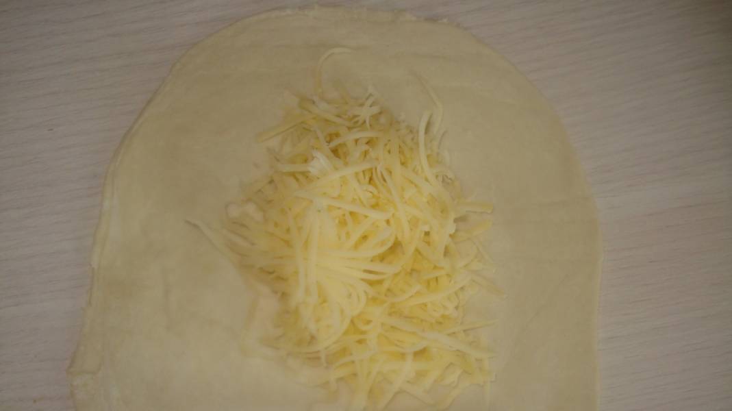 1. Тесто размораживаем, а сыр трем на терке. Делим тесто на одинаковые кусочки. Каждый кусочек раскатываем в пласт, по центру которого выкладываем тертый сыр. 
