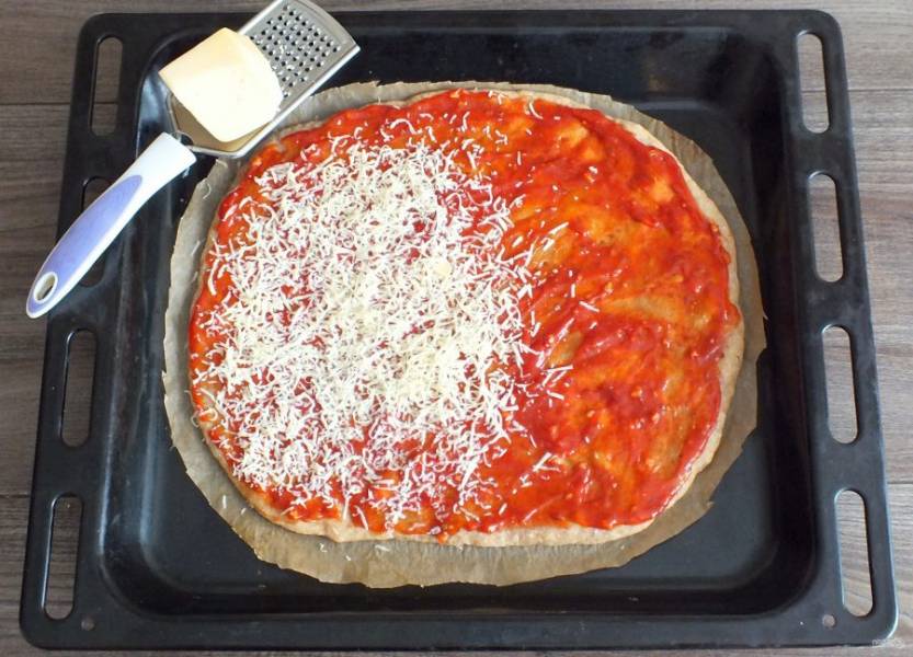 Через указанное время достаньте противень с пластом из духовки. Духовку не выключайте. Основу смажьте томатным соусом. Сверху натрите 80 грамм сыра.