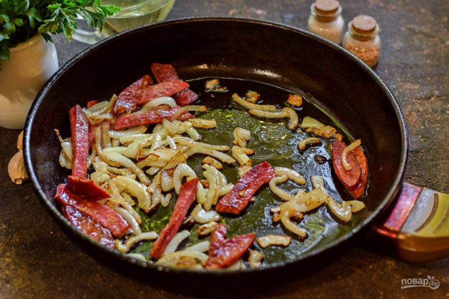 В сковороде прогрейте растительное масло, выложите лук и колбасу. Поджарьте ингредиенты 3-4 минуты.