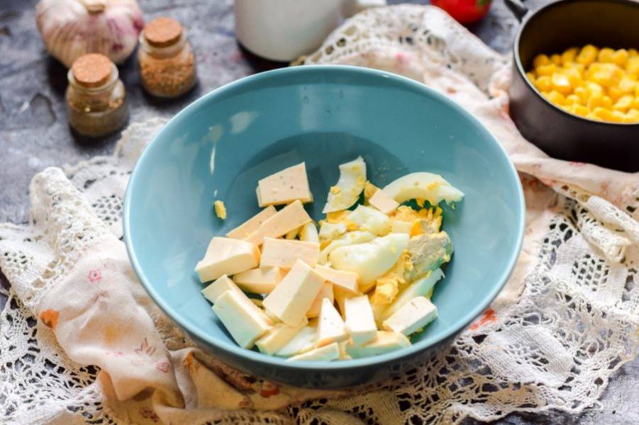Куриное яйцо отварите заранее и остудите, нарежьте небольшими кусочками и переложите в салатник. Плавленый сыр возьмите с зеленью или со вкусом бекона, нарежьте сыр кусочками и добавьте к яйцу.