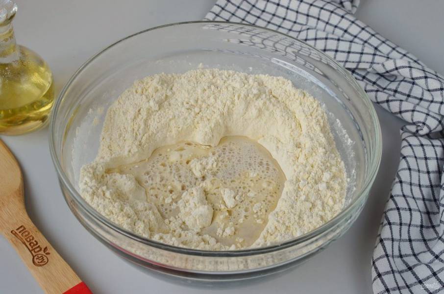 В муке сделайте ямку по центру, влейте воду с дрожжами, ложкой начните замешивать тесто, в процессе добавляя растительное масло.